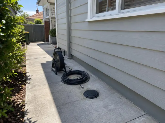 Capped drain inspection opening beside a coiled camera reel on a concrete side path by a weatherboard house in Mascot