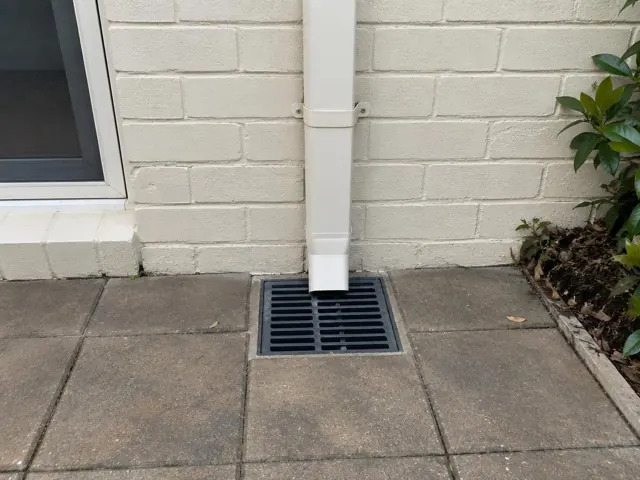 Clean stormwater grate beneath a white downpipe beside a brick house and paved patio in Eastlakes